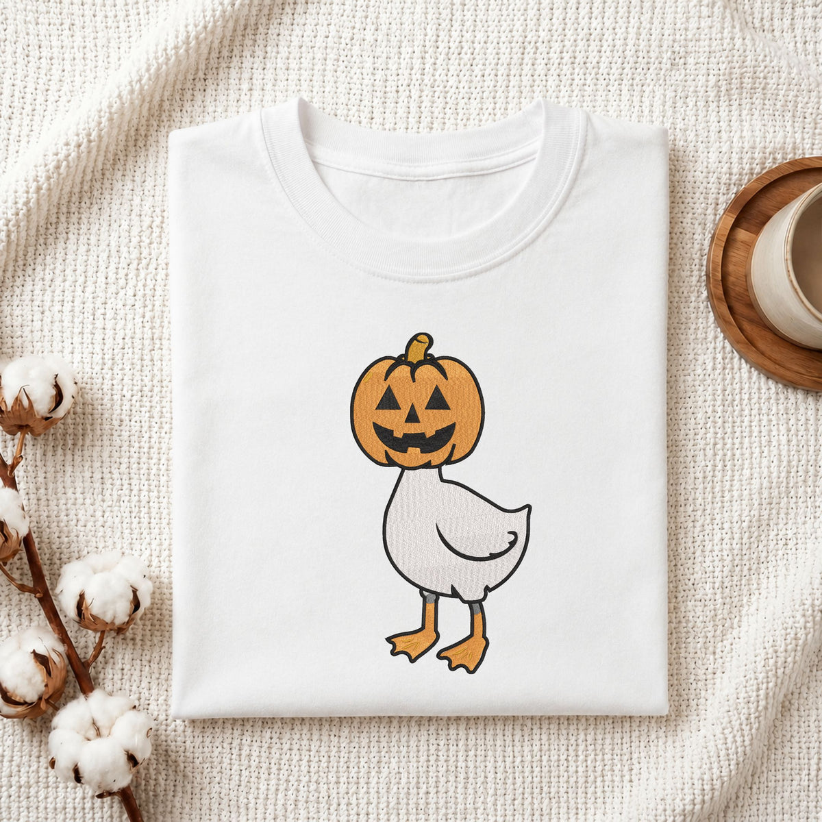 Folded white cotton t-shirt mockup featuring a cute duck in a pumpkin head Halloween machine embroidery design, styled with cotton stems and a wooden cup