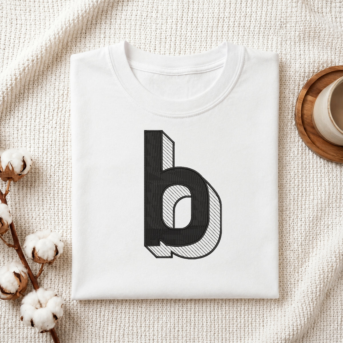 A neatly folded white crewneck t-shirt featuring a stitched 3D block lowercase letter "b" design, styled with cotton branches and a wooden coaster.