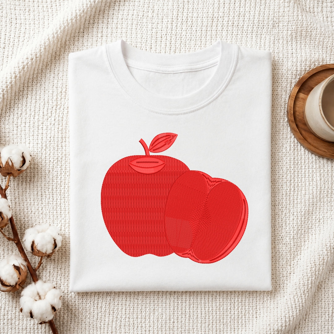 A folded white crewneck t-shirt featuring a textured red apple graphic on the front, styled on a beige woven blanket with cotton bolls and a wooden cup in the background.