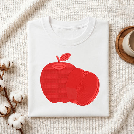 A folded white crewneck t-shirt featuring a textured red apple graphic on the front, styled on a beige woven blanket with cotton bolls and a wooden cup in the background.