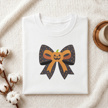 A neatly folded white t-shirt featuring the embroidered pumpkin bow design, displayed on a textured white knit background with cotton stems and a wooden cup.