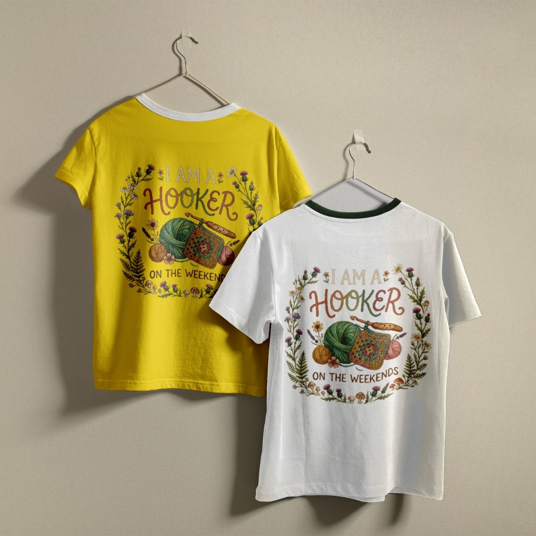 Two children's short-sleeve ringer T-shirts hanging on hangers against a plain beige wall. One shirt is bright yellow and the other is white, both featuring a large front graphic with the words 'I AM A HOOKER ON THE WEEKENDS' in a floral wreath design around a green turtle and a peach.