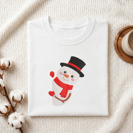  Cute snowman wearing a black top hat, red band, and red scarf machine embroidery design displayed in a wooden hoop on white fabric with thread spools and a needle.