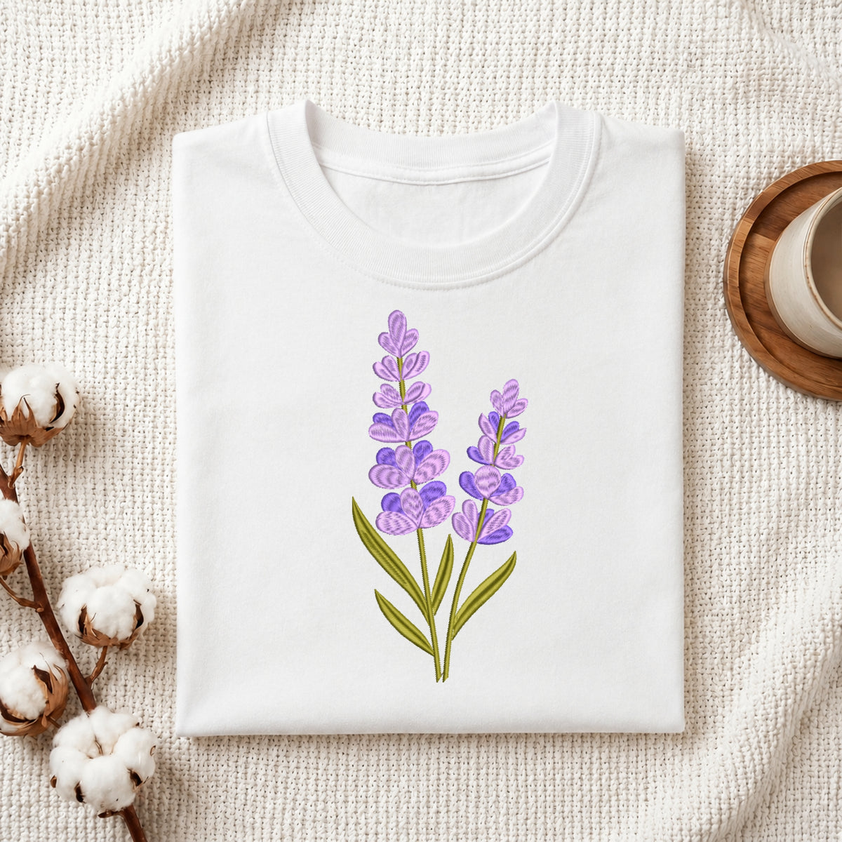 A folded white crewneck t-shirt featuring a watercolor-style lavender illustration on the front, styled on a textured cream knit blanket with cotton bolls and a wooden cup in the background.