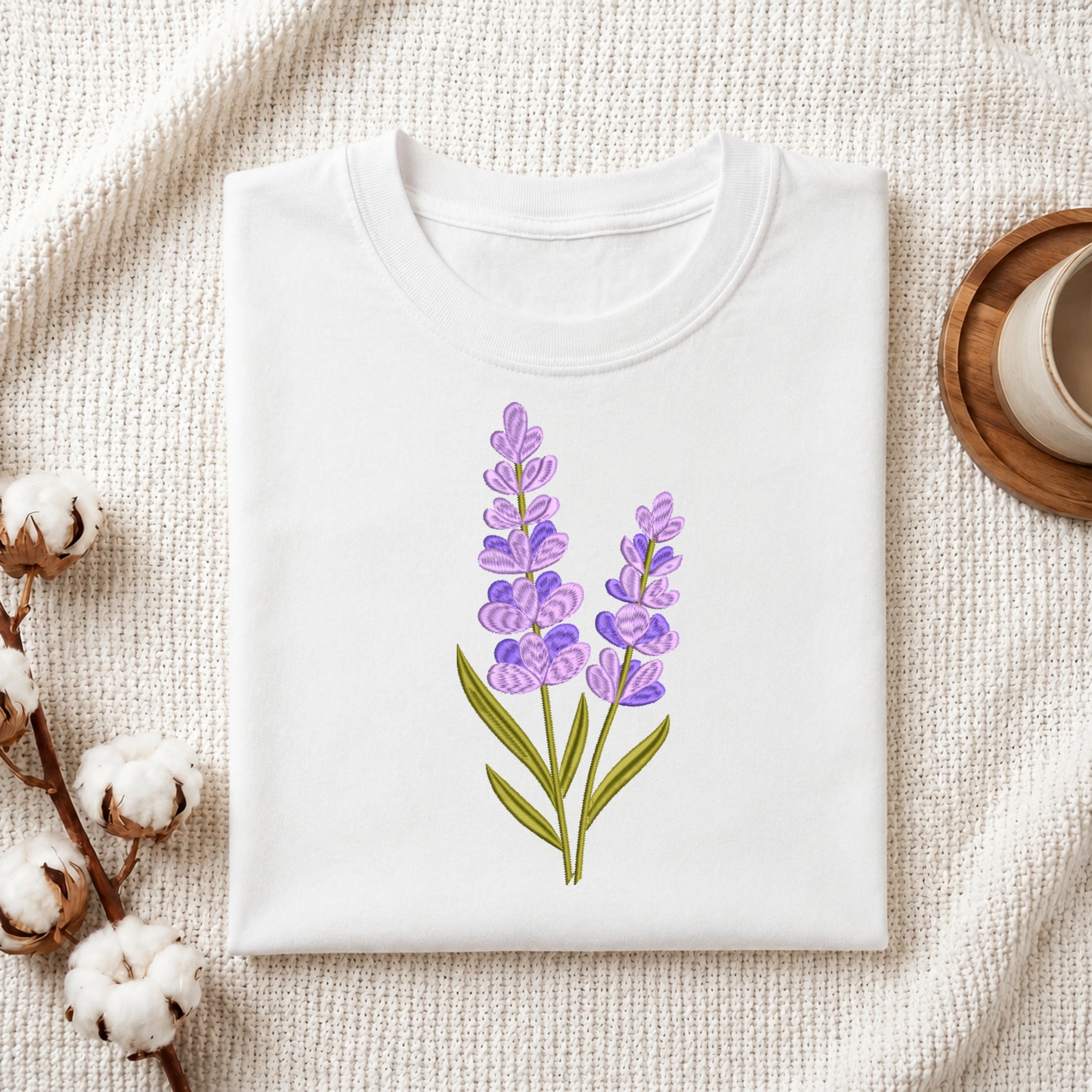 A folded white crewneck t-shirt featuring a watercolor-style lavender illustration on the front, styled on a textured cream knit blanket with cotton bolls and a wooden cup in the background.
