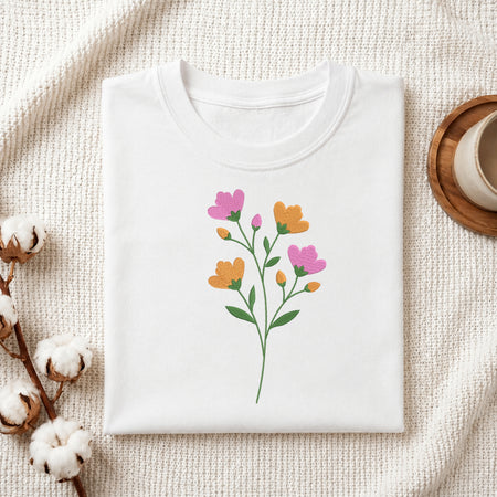 A folded white crewneck t-shirt with a colorful floral graphic print, styled on a textured cream knit blanket with cotton bolls and a wooden cup in the background.