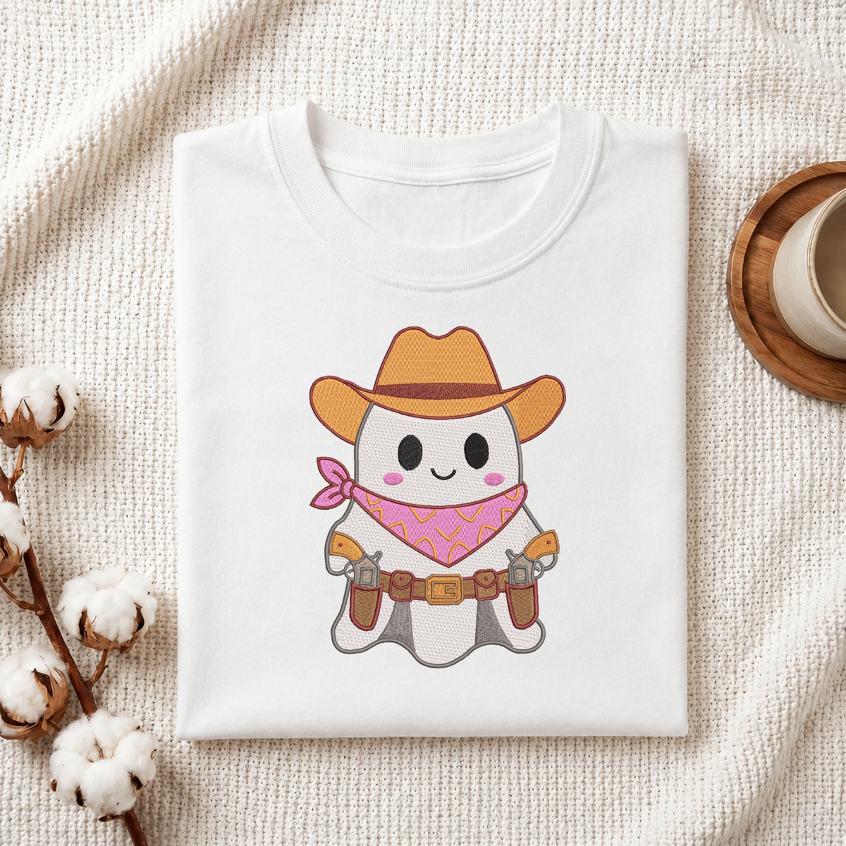 A neatly folded white t-shirt featuring the embroidered cowboy ghost design, displayed on a textured white knit background with cotton stems and a wooden cup.