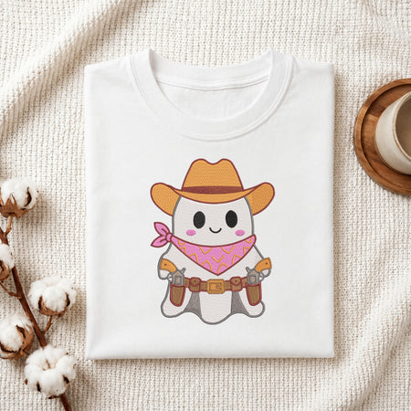 A neatly folded white t-shirt featuring the embroidered cowboy ghost design, displayed on a textured white knit background with cotton stems and a wooden cup.