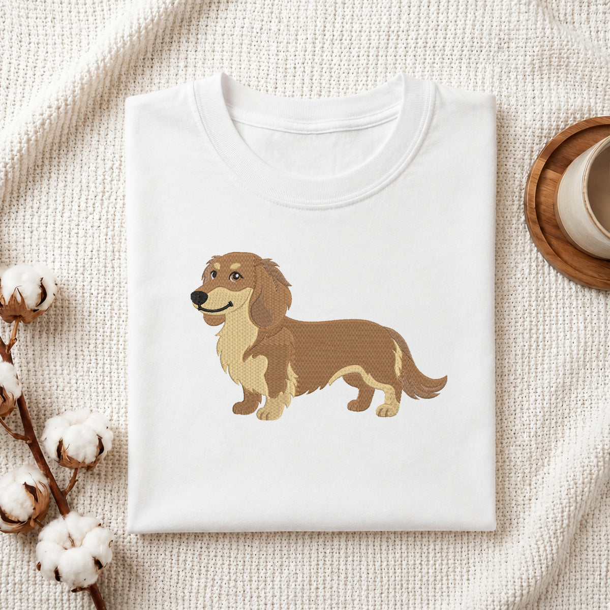 A neatly folded white crewneck t-shirt featuring a stitched cute dachshund dog design, styled with cotton branches and a wooden coaster.
