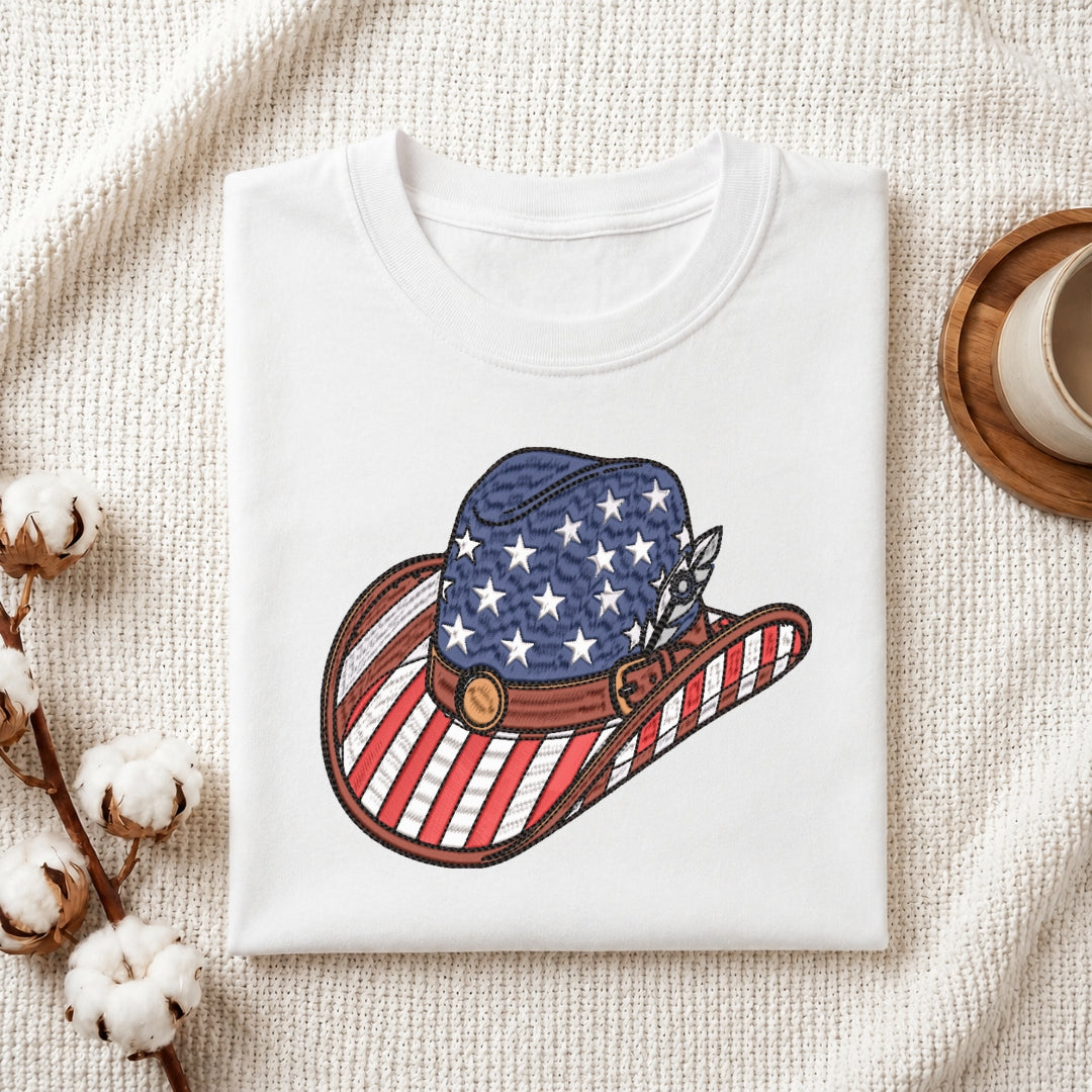 A folded white crewneck t-shirt featuring a graphic of a cowboy hat decorated with an American flag pattern (blue with white stars on the crown, red and white stripes on the brim and side, with a brown leather-look band and silver feather). The t-shirt is styled in a flat-lay on a textured cream knit blanket, with cotton bolls and a wooden cup visible as props.