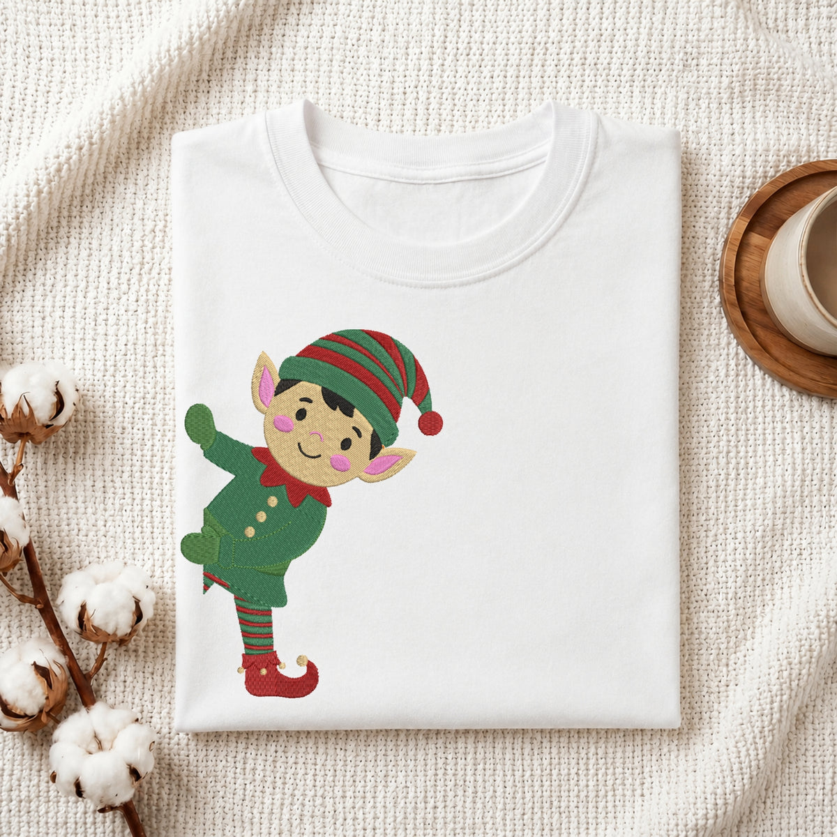 A folded white crewneck t-shirt featuring a cartoon elf graphic, styled on a textured cream knit blanket with cotton bolls and a wooden cup in the background.