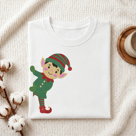 A folded white crewneck t-shirt featuring a cartoon elf graphic, styled on a textured cream knit blanket with cotton bolls and a wooden cup in the background.