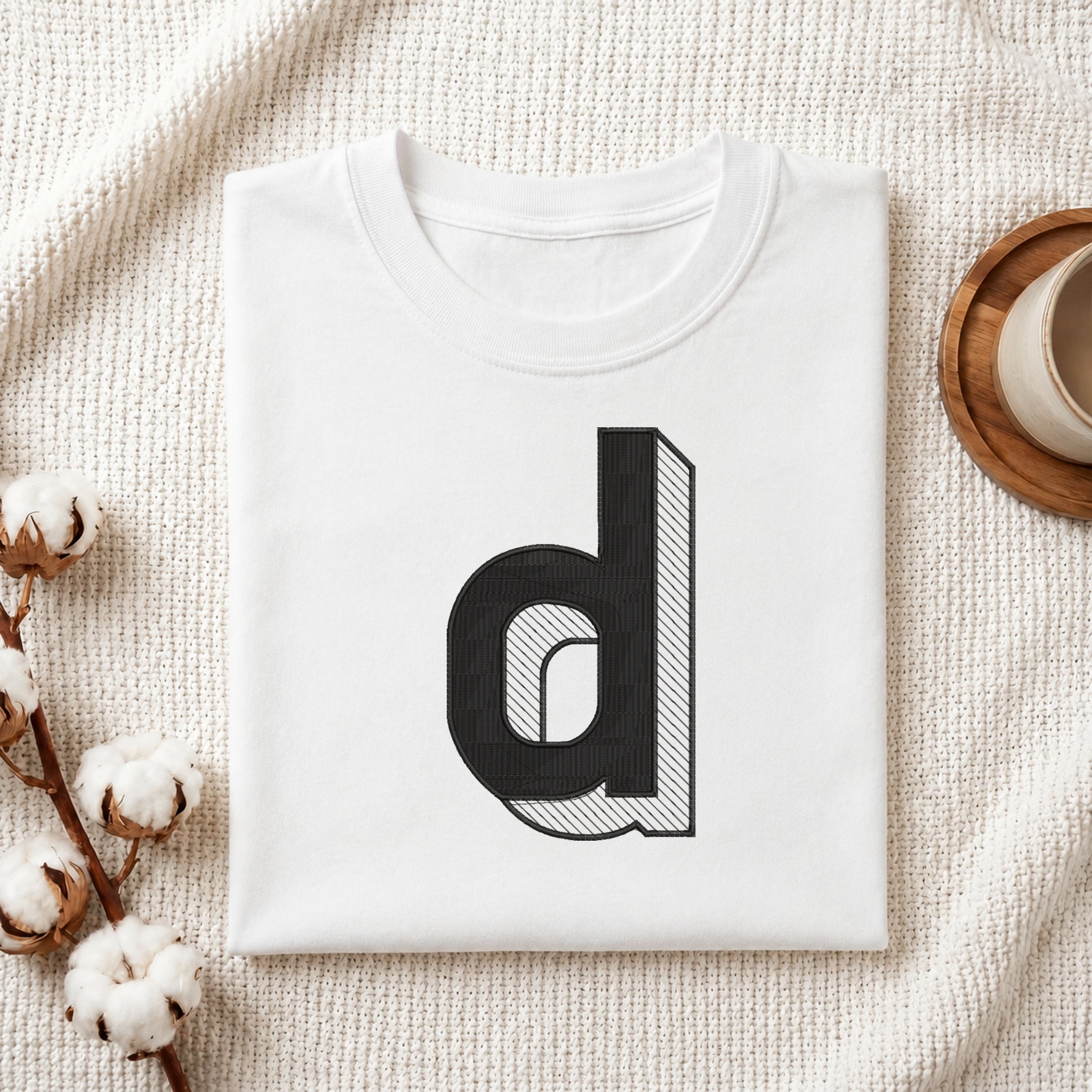 A folded white t-shirt with a large black lowercase letter 'd' graphic printed on the front, styled on a textured cream knit blanket with cotton bolls and a wooden cup in the background.