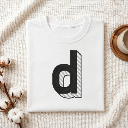 A folded white t-shirt with a large black lowercase letter 'd' graphic printed on the front, styled on a textured cream knit blanket with cotton bolls and a wooden cup in the background.