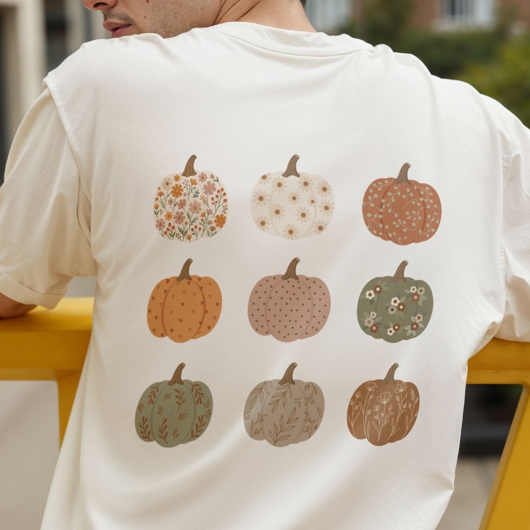 A person wearing a cream-colored t-shirt, shown from the back. The shirt features a graphic print of nine autumn pumpkins in various patterns and muted fall colors, including orange, brown, green, and beige, arranged in a 3x3 grid.