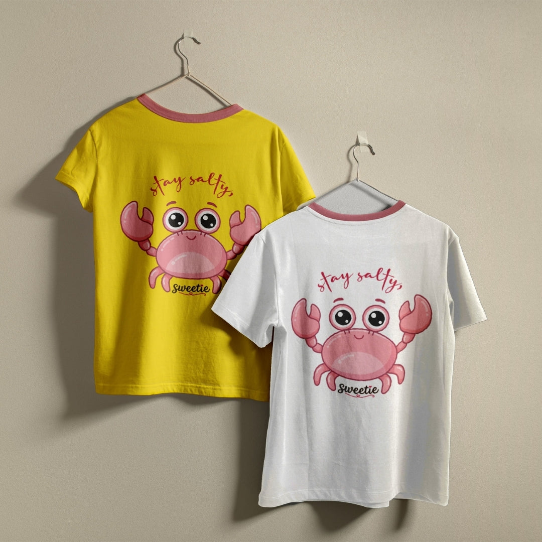 Two t-shirts hanging on hangers, one bright yellow and one white, both printed with a cartoon pink crab graphic and the text "stay salty, Sweetie".