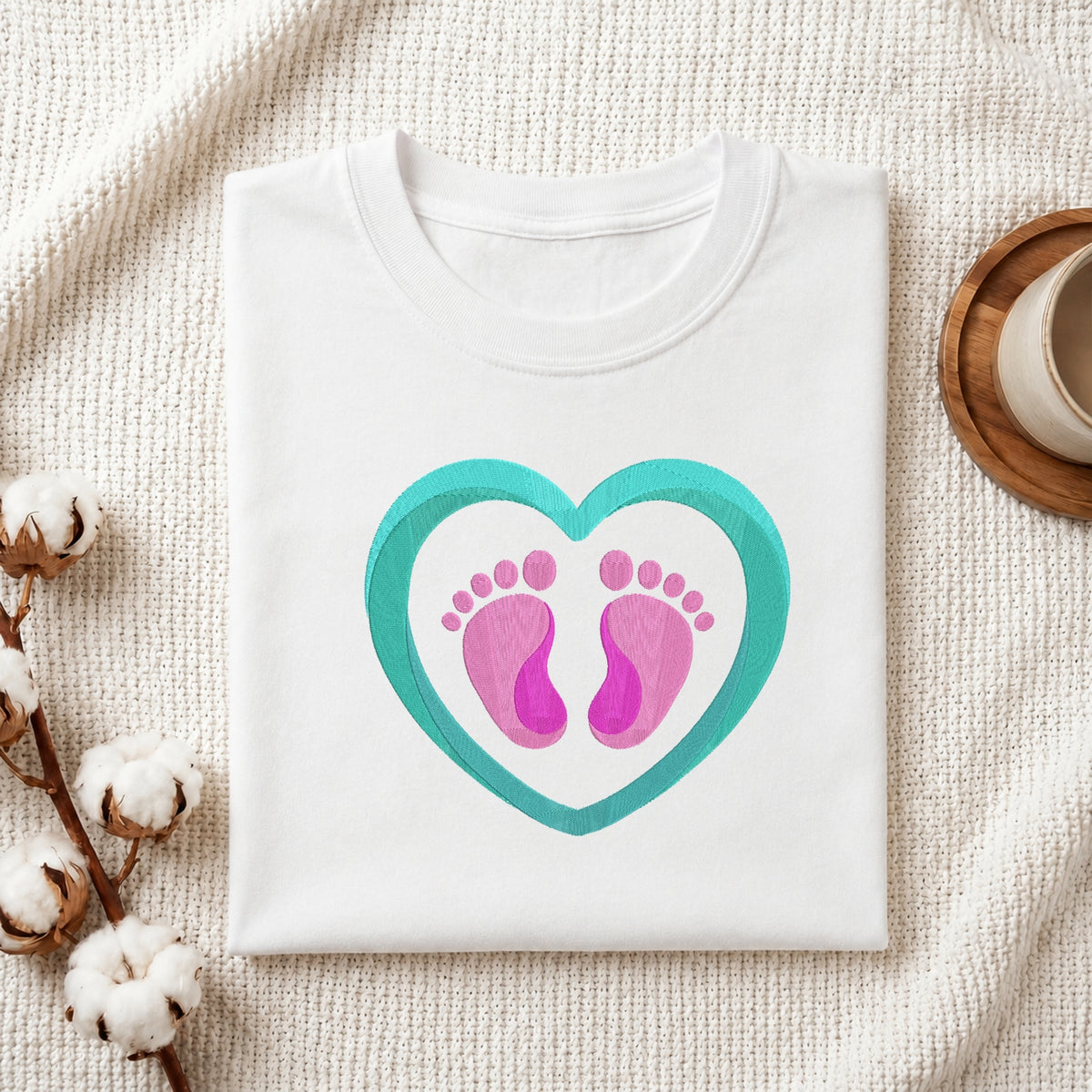 Folded white cotton t-shirt mockup featuring a sweet baby footprint and heart machine embroidery design, styled with cotton stems and a wooden cup.