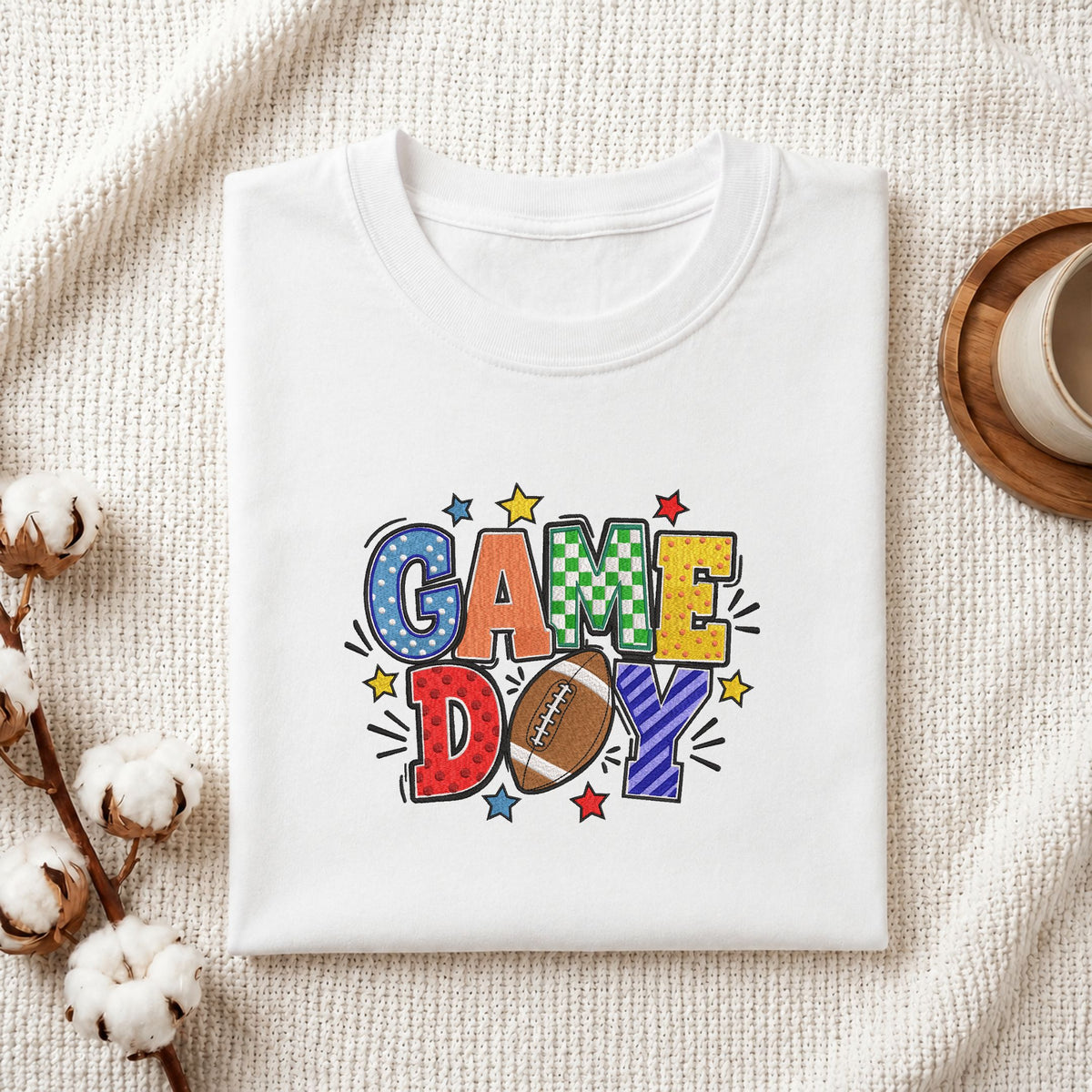 Folded white cotton t-shirt mockup featuring a "Game Day" football machine embroidery design, styled with cotton stems and a wooden cup   