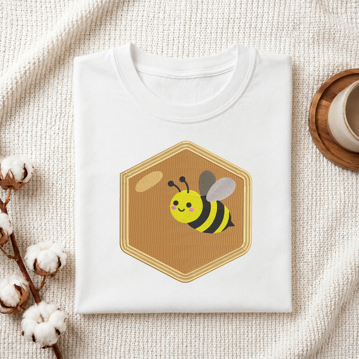 A folded white T-shirt with a cartoon bee graphic inside a tan honeycomb shape, styled on a textured cream fabric with cotton stems and a small cup in the background.