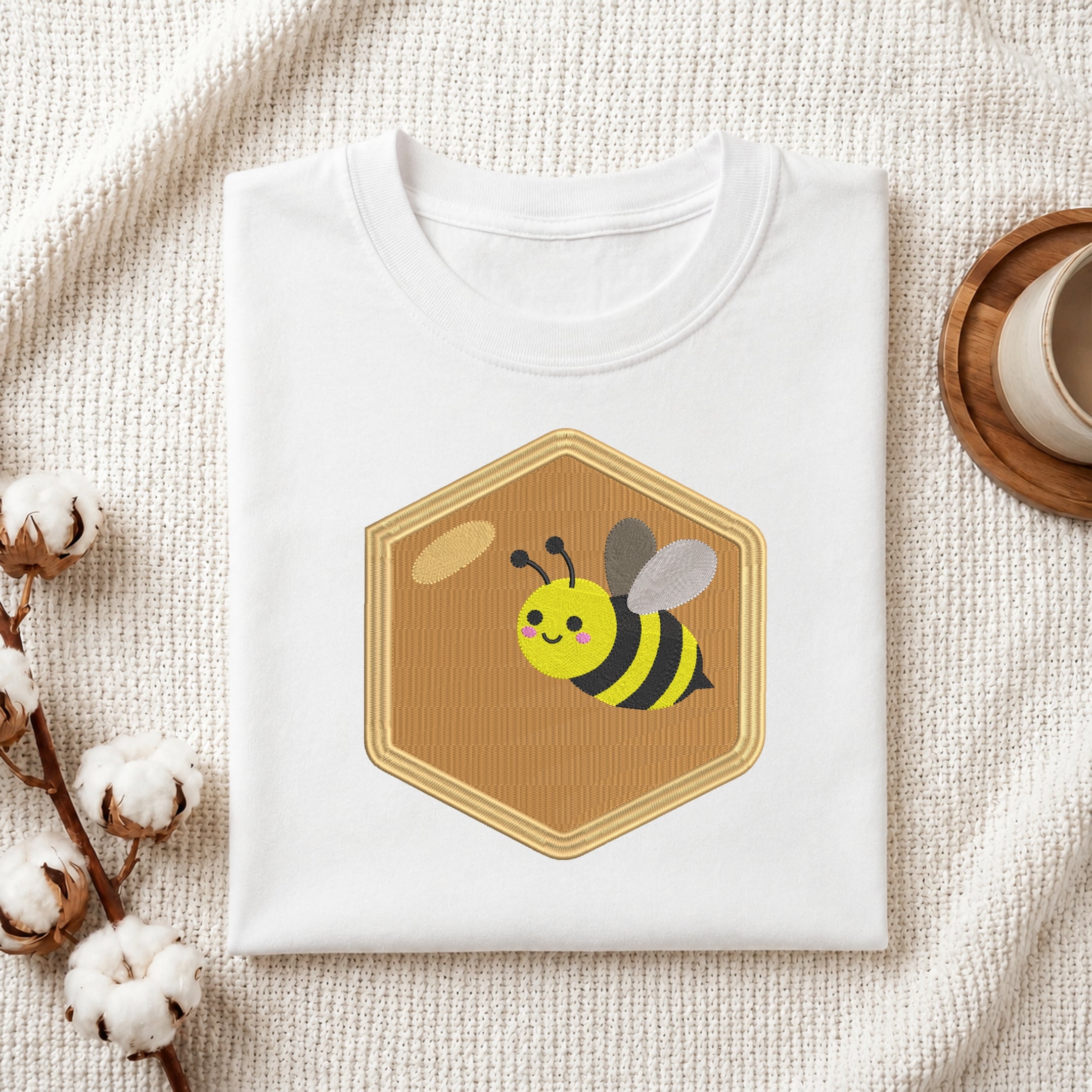 A folded white T-shirt with a cartoon bee graphic inside a tan honeycomb shape, styled on a textured cream fabric with cotton stems and a small cup in the background.