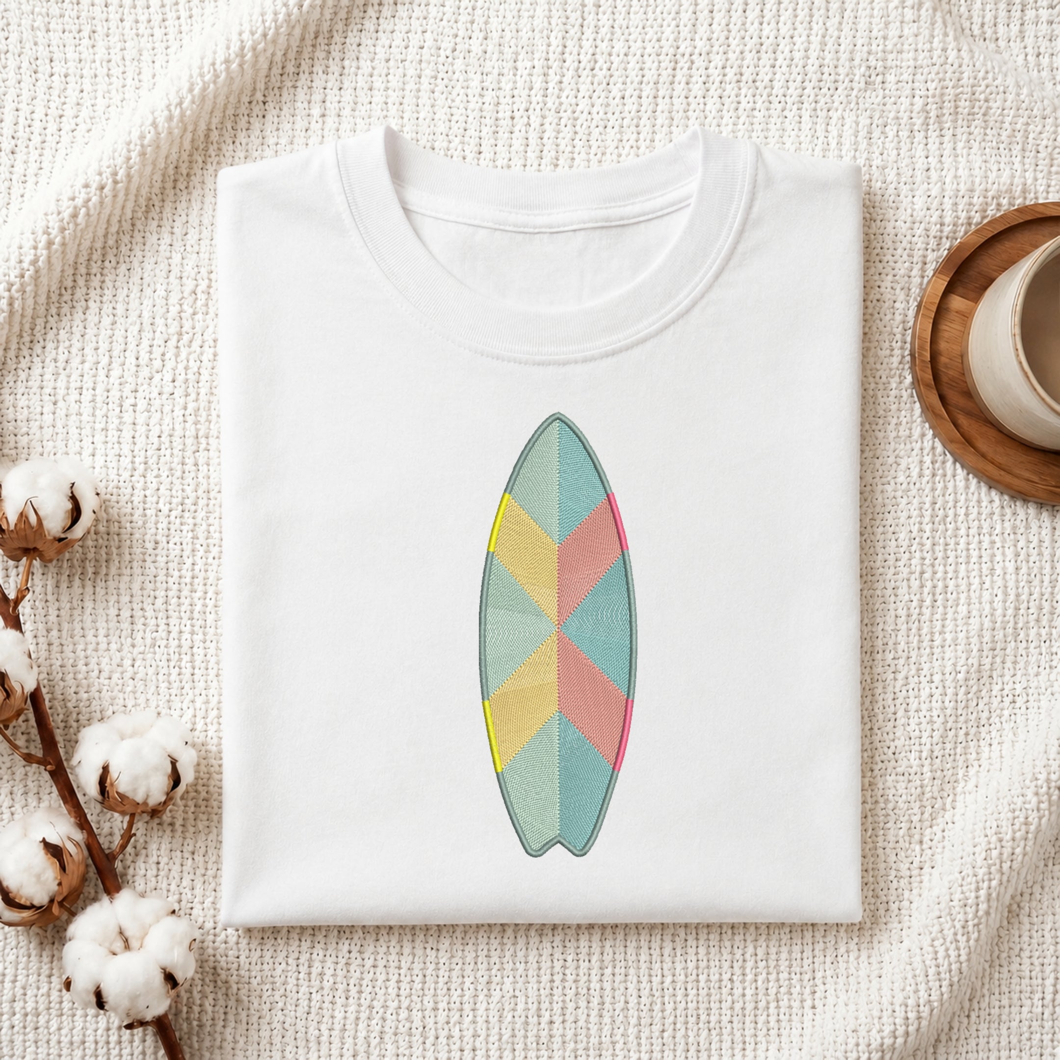 A folded white crewneck t-shirt featuring a colorful geometric surfboard graphic on the chest, styled on a textured cream fabric with cotton bolls and a wooden cup.
