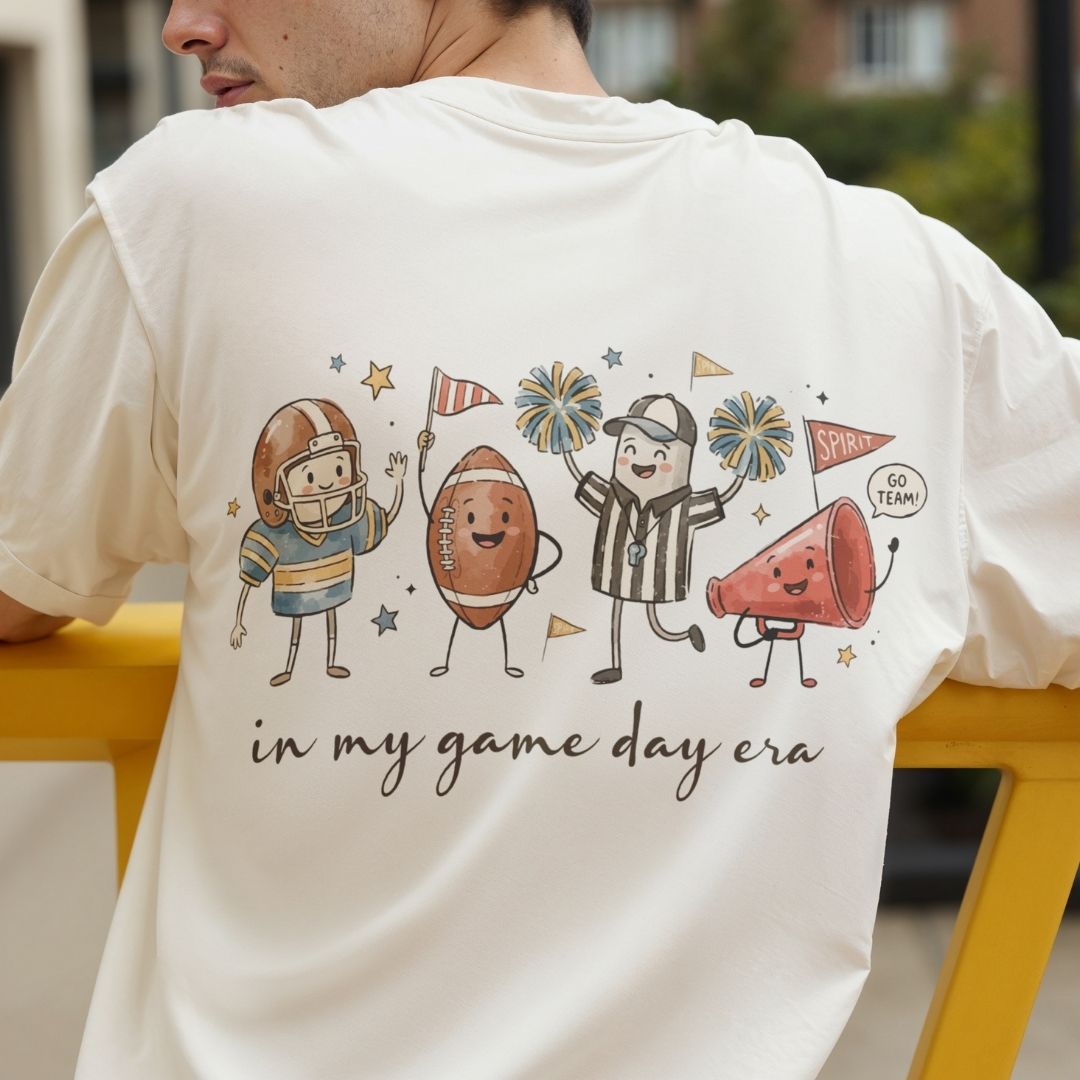 A man wearing a cream-colored t-shirt, showing the back of the shirt with a colorful cartoon graphic of sports figures and the text 'in my game day era'.