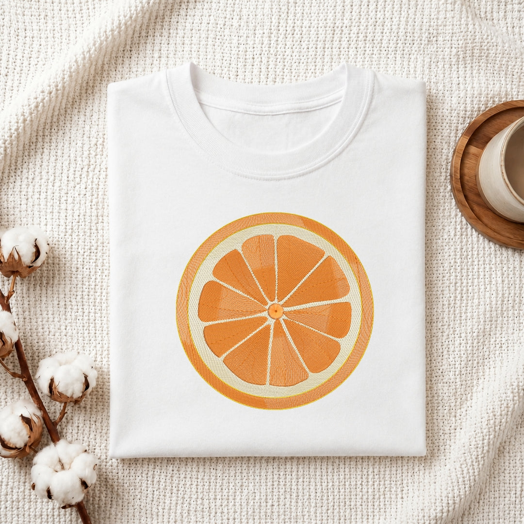 A folded white crewneck t-shirt featuring a large graphic print of a sliced orange on the front, styled on a textured cream blanket with cotton bolls and a wooden cup in the background.