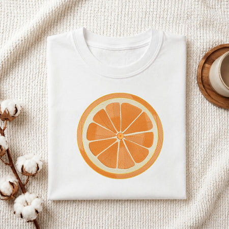 A folded white crewneck t-shirt featuring a large graphic print of a sliced orange on the front, styled on a textured cream blanket with cotton bolls and a wooden cup in the background.