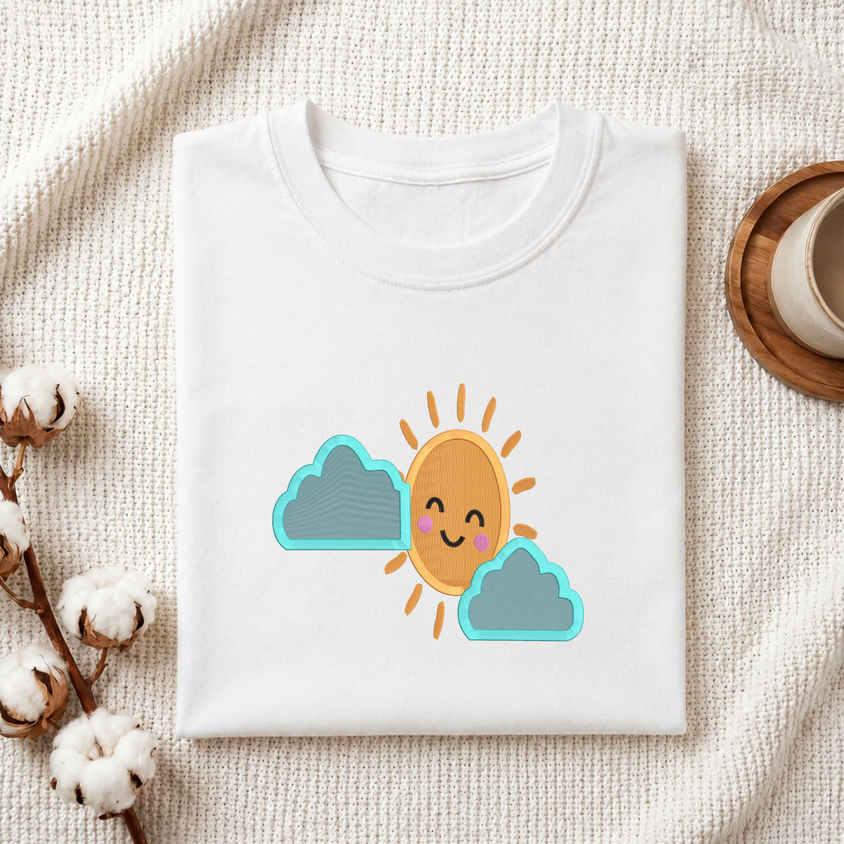 Folded white cotton t-shirt mockup featuring a cute sun and clouds machine embroidery design, styled with cotton stems and a wooden cup