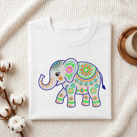 A folded white crewneck t-shirt featuring a colorful, hand-drawn style elephant illustration with purple, green, orange, and pink patterns, styled on a textured cream fabric with cotton bolls and a wooden cup.