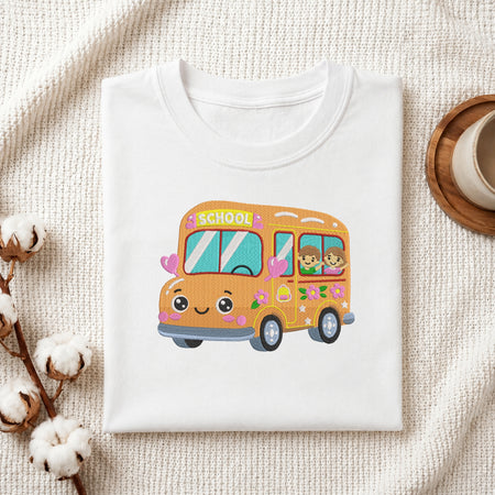 A folded white T-shirt displayed on a textured cream fabric background, featuring a cute cartoon school bus graphic in orange, pink, yellow, blue, and green with the word 'SCHOOL' on the front.