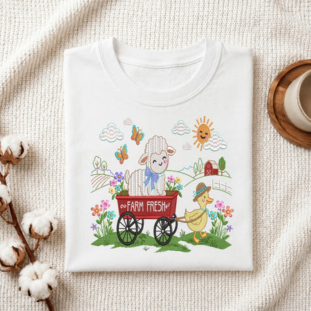 A folded white crewneck t-shirt with a colorful cartoon graphic of a sheep in a wagon and a duck, styled on a textured cream fabric with cotton bolls and a coffee cup.