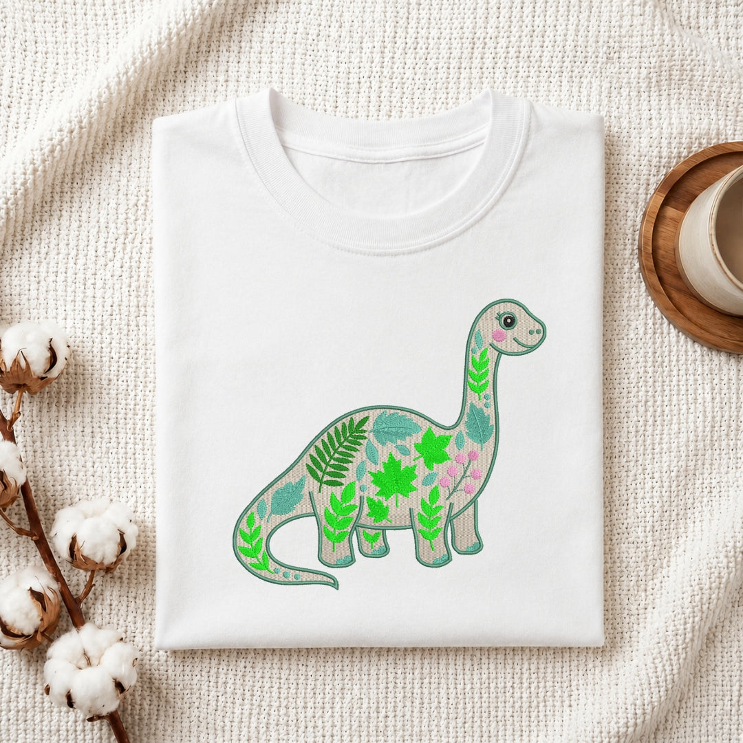 A folded white crewneck t-shirt displayed on a textured cream linen background, decorated with cotton bolls on the left and a wooden cup on the right. The t-shirt features a graphic of a long-necked dinosaur filled with green leaves and pink floral patterns.