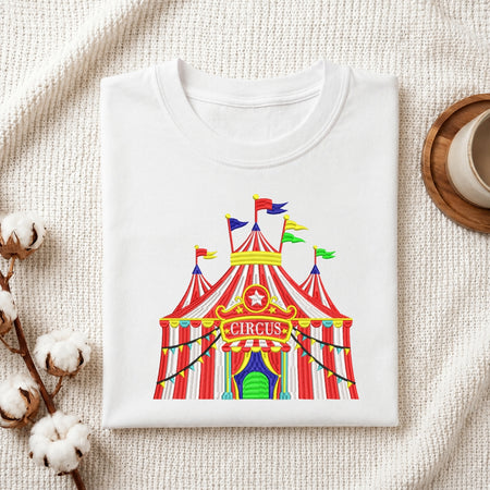 A folded white T-shirt with a colorful circus tent graphic on the front, styled on a textured cream fabric with cotton stems and a small wooden cup in the background.