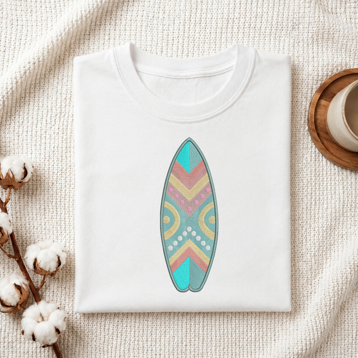 A folded white crewneck t-shirt featuring a colorful retro-patterned surfboard graphic on the front, styled on a textured cream fabric with cotton bolls and a wooden cup.