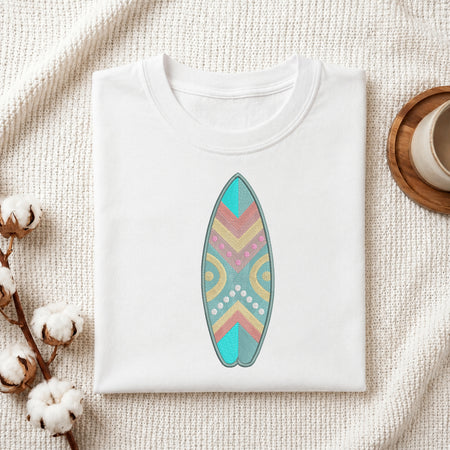 A folded white crewneck t-shirt featuring a colorful retro-patterned surfboard graphic on the front, styled on a textured cream fabric with cotton bolls and a wooden cup.