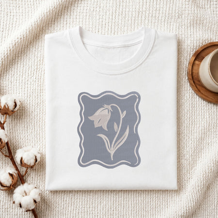 Folded white cotton t-shirt mockup featuring a purple bellflower machine embroidery design, styled with cotton stems and a wooden cup.