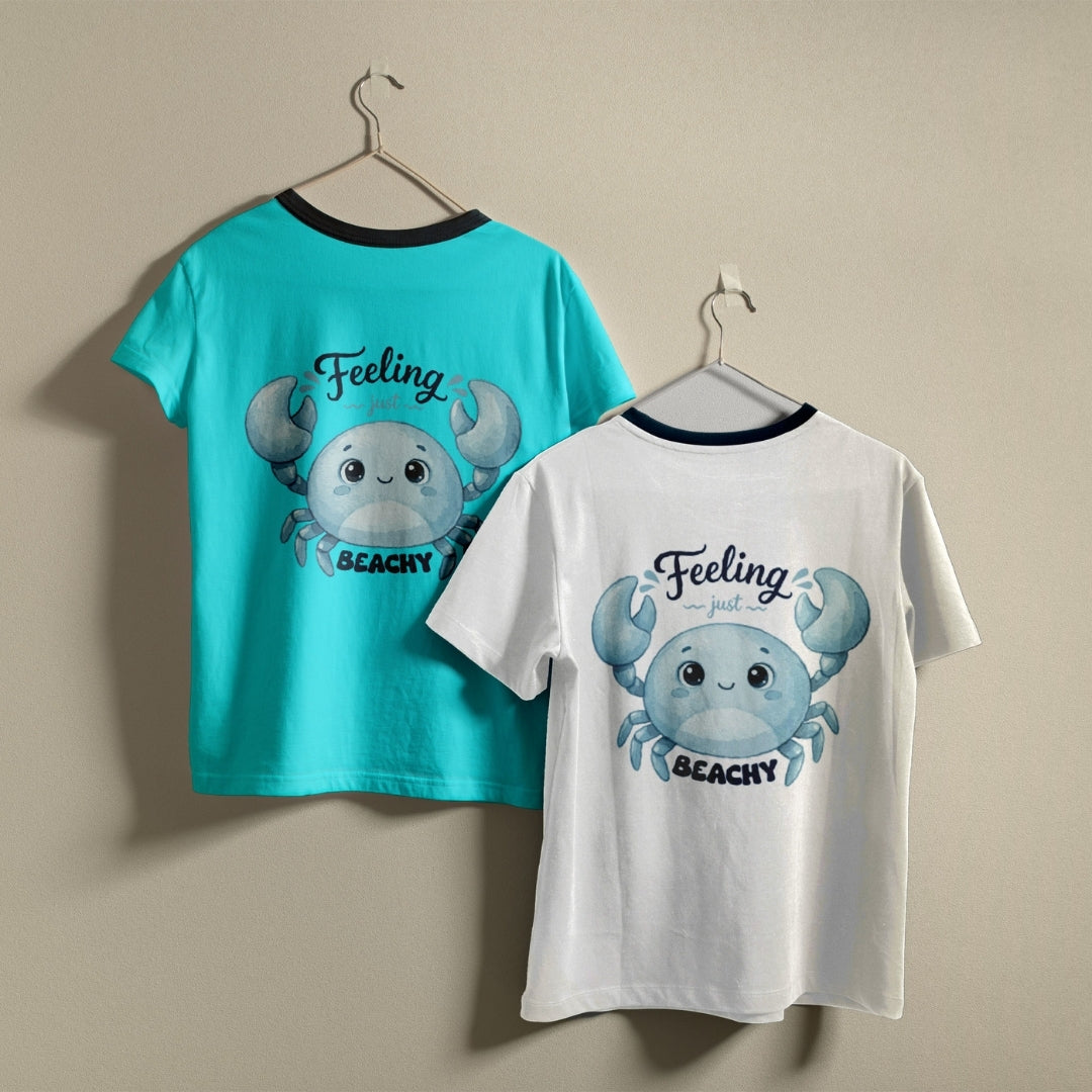 Two t-shirts hanging on hangers, one turquoise and one white, both printed with a blue cartoon crab graphic and the text "Feeling just BEACHY".