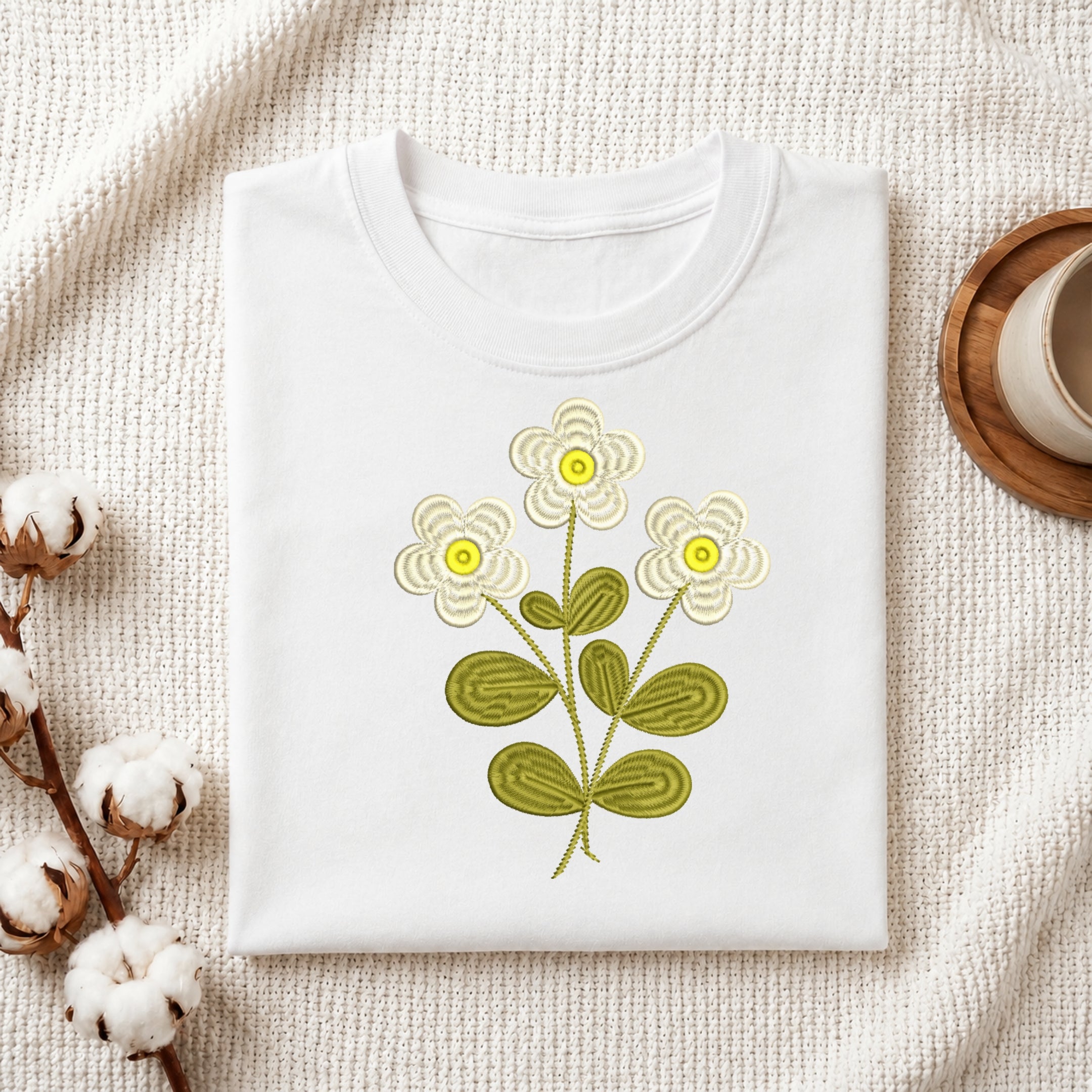 A folded white T-shirt with a stylized embroidered floral design featuring three cream flowers with yellow centers and olive green leaves, displayed on a textured cream fabric with cotton bolls and a wooden cup in the background.