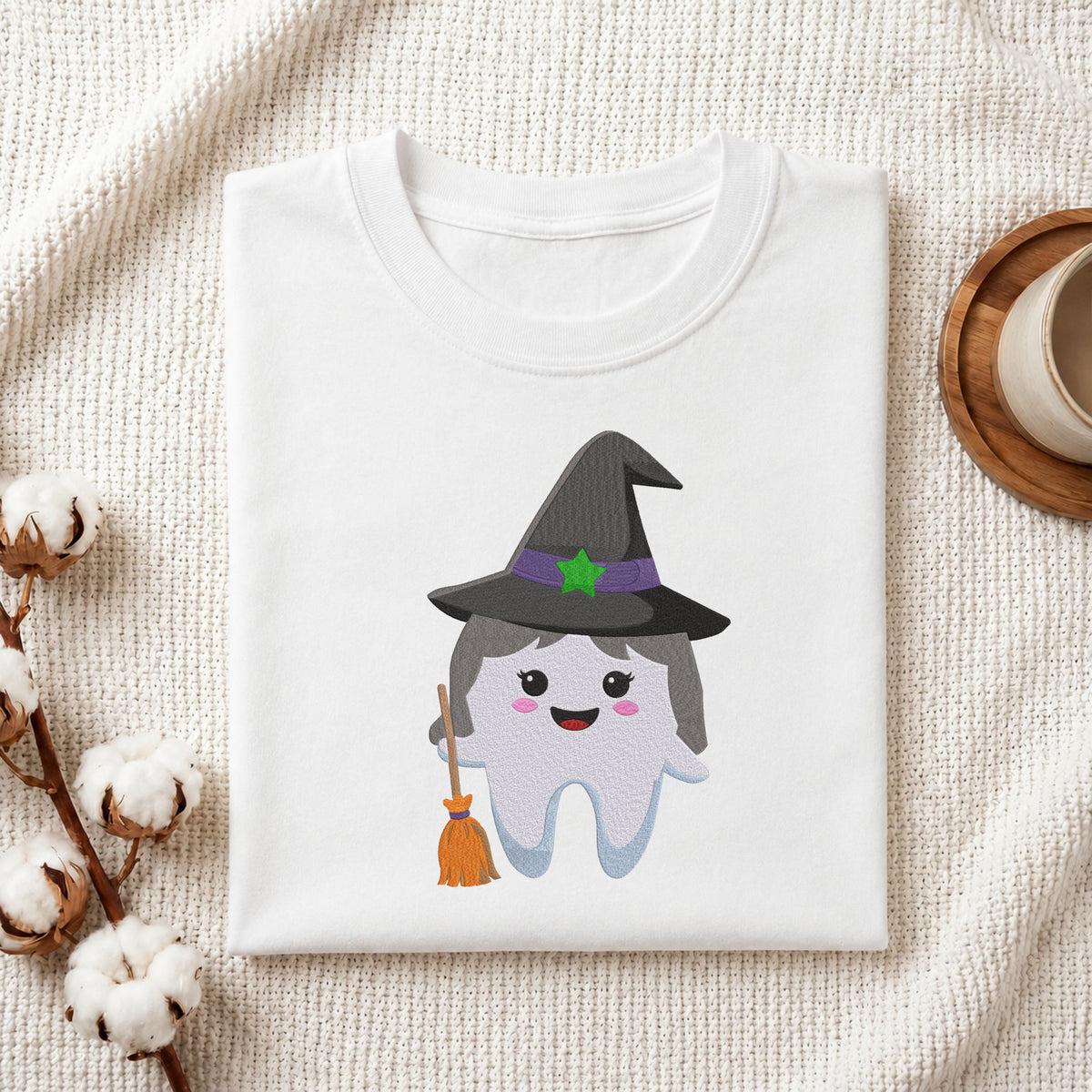Folded white cotton t-shirt mockup featuring a cute witch tooth Halloween dental machine embroidery design, styled with cotton stems and a wooden cup