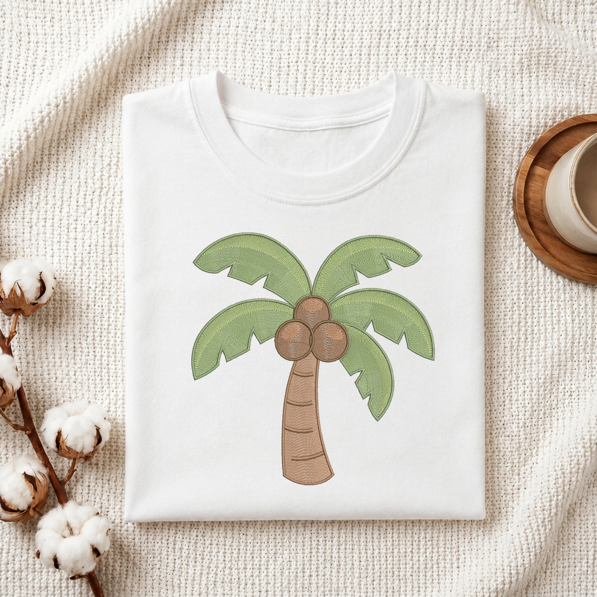 A folded white T-shirt with a cartoon palm tree graphic featuring green leaves and brown coconuts, styled on a textured cream fabric with cotton bolls and a wooden cup in the background.