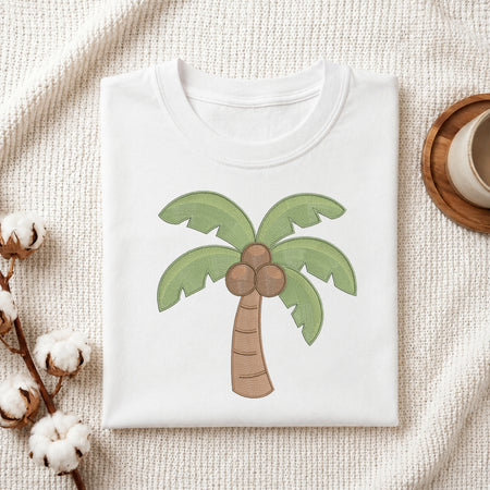 A folded white T-shirt with a cartoon palm tree graphic featuring green leaves and brown coconuts, styled on a textured cream fabric with cotton bolls and a wooden cup in the background.