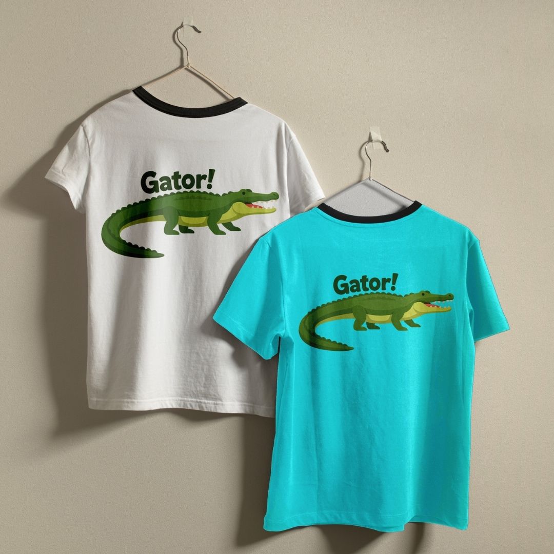 Two t-shirts with a black ringer collar—one white and one vibrant teal—hanging on a neutral wall, both showcasing a green cartoon alligator design.