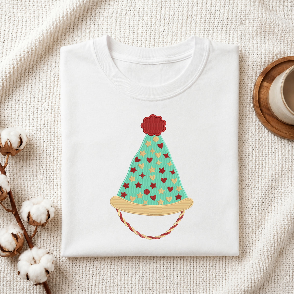 A folded white crewneck t-shirt featuring a colorful embroidered or printed party hat design, styled on a textured cream fabric with cotton bolls and a wooden cup in the background.