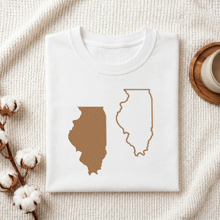 A folded white T-shirt laid flat on a textured cream fabric background, featuring a brown filled silhouette of Illinois and a matching outline on the front. Cotton stems and part of a wooden cup are visible as styled props.