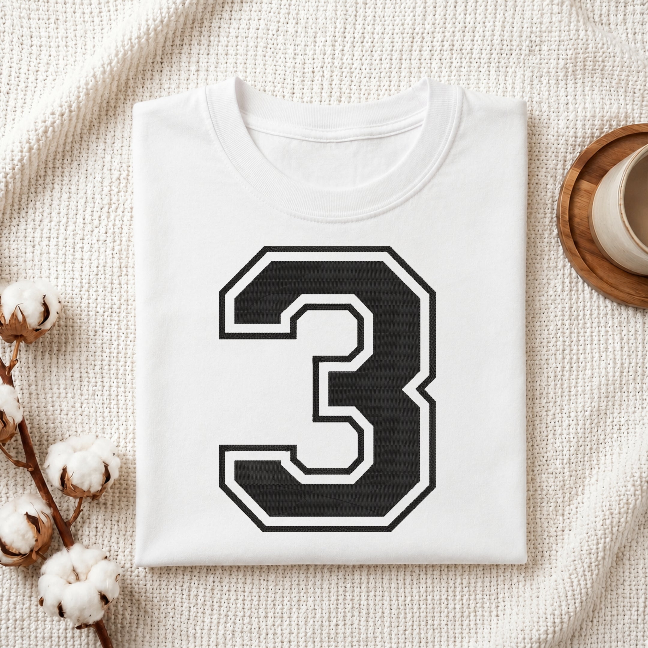 A folded white T-shirt with a large black number 3 outlined in white on the front, styled on a textured cream fabric with cotton stems and a small cup in the background.