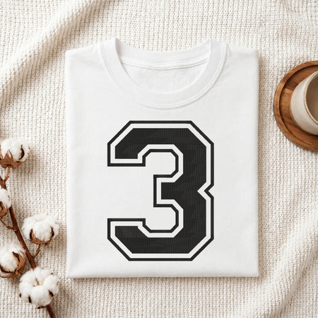 A folded white T-shirt with a large black number 3 outlined in white on the front, styled on a textured cream fabric with cotton stems and a small cup in the background.