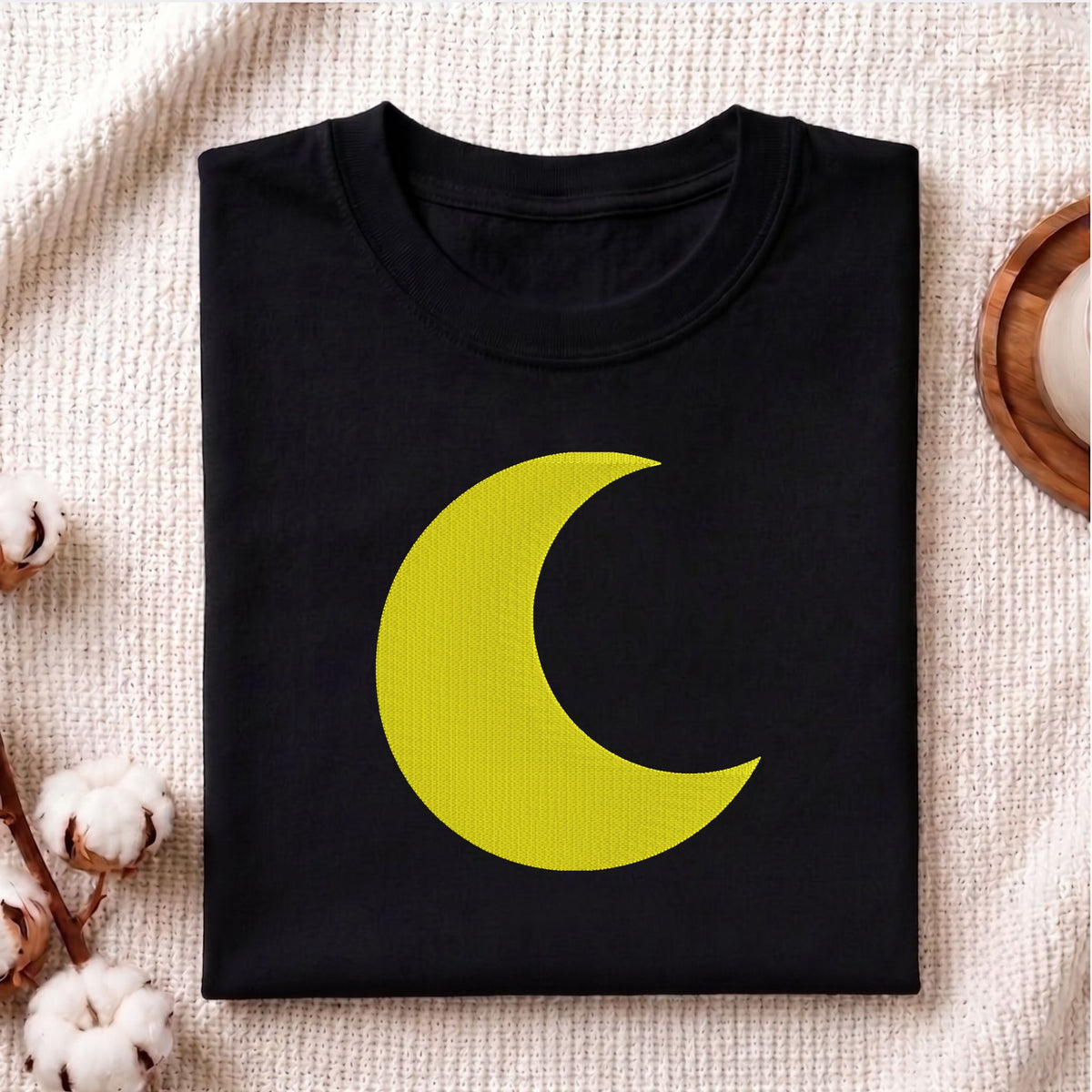 A folded black crewneck t-shirt featuring a large yellow crescent moon graphic on the front, styled on a textured cream knit fabric with cotton bolls and a wooden tray.