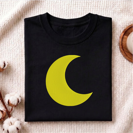 A folded black crewneck t-shirt featuring a large yellow crescent moon graphic on the front, styled on a textured cream knit fabric with cotton bolls and a wooden tray.