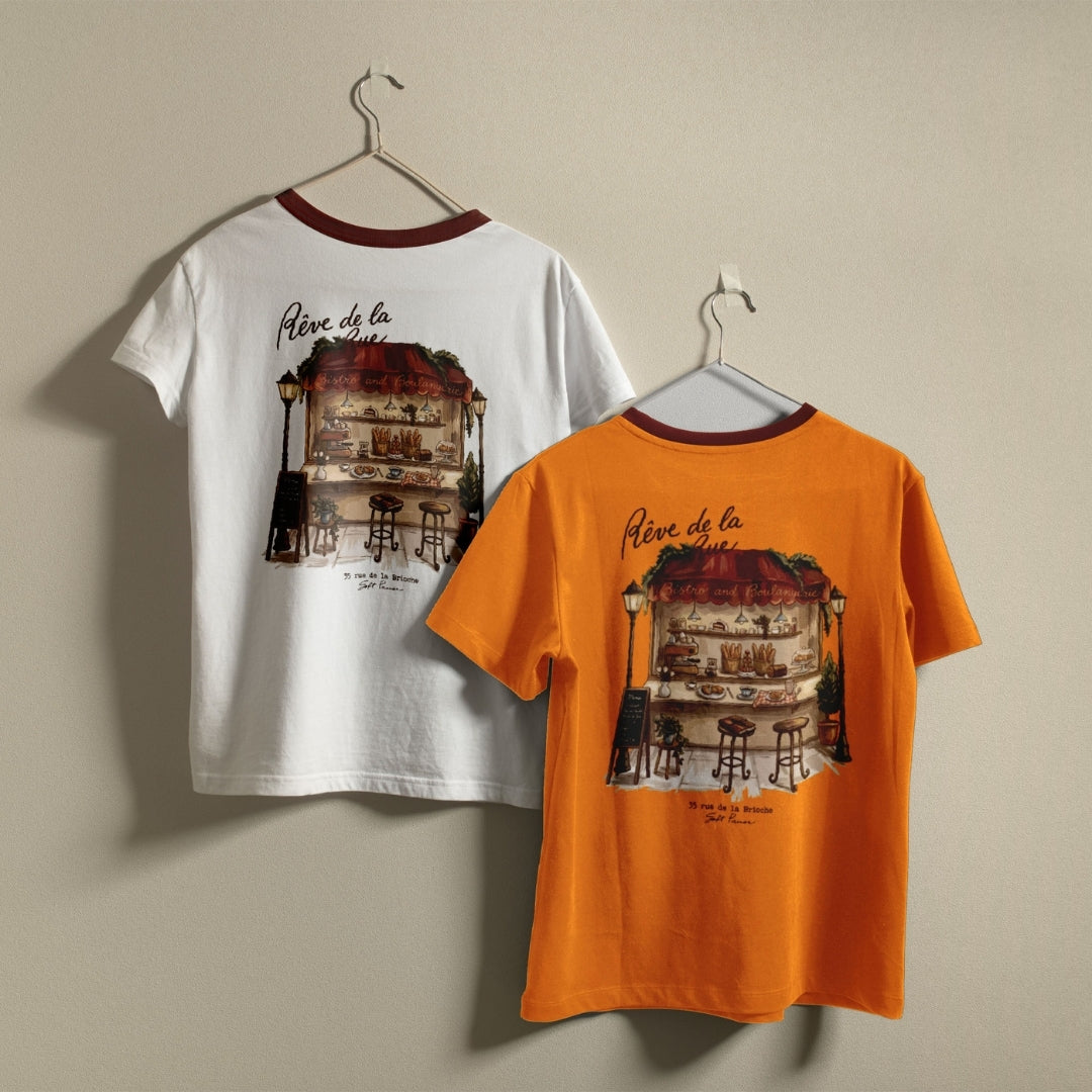 White and orange t-shirts hanging on a wall, both displaying the intricate French cafe and boulangerie graphic printed on the back.
